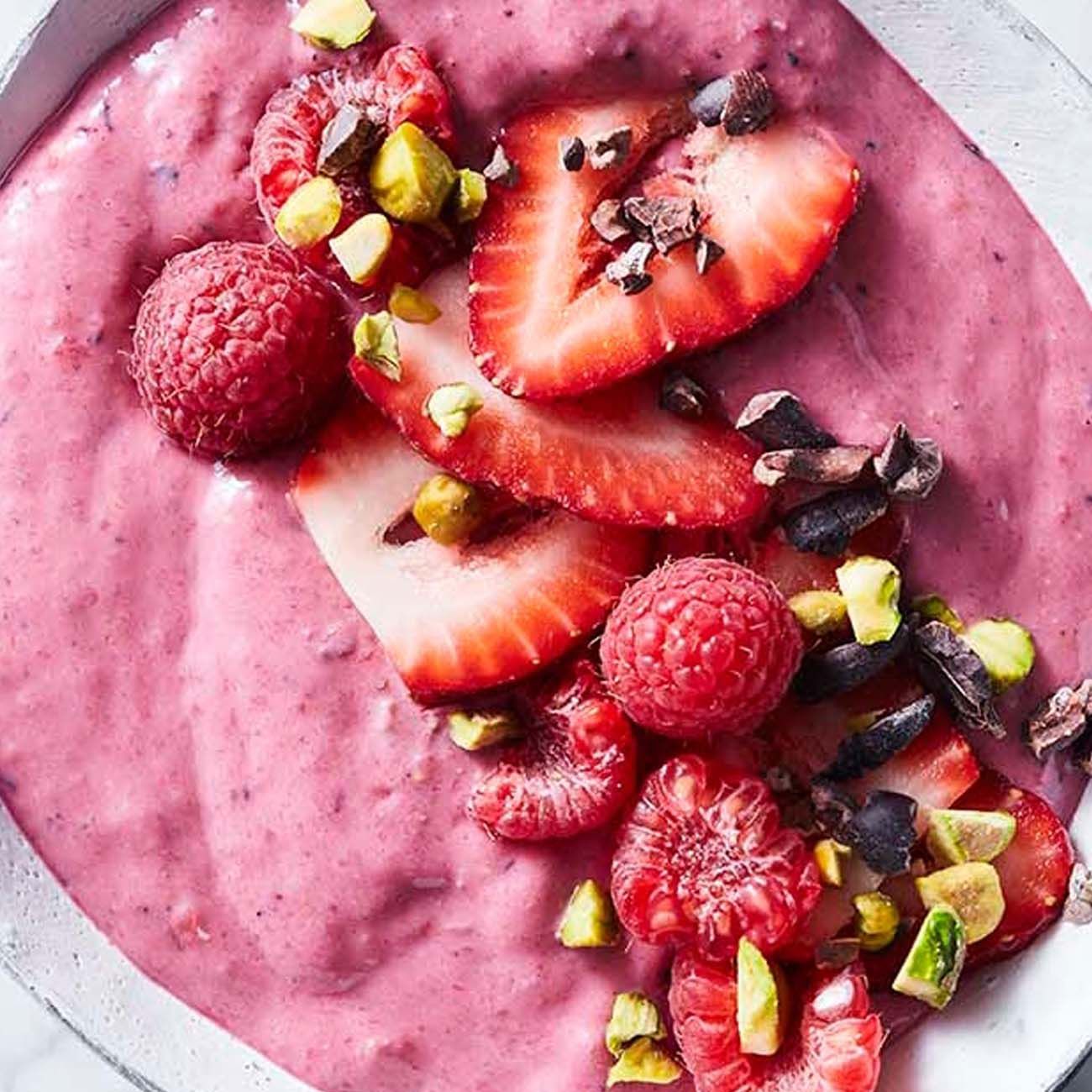 A close-up of a pink berry smoothie bowl topped with sliced strawberries, whole raspberries, chopped pistachios and cacao nibs.