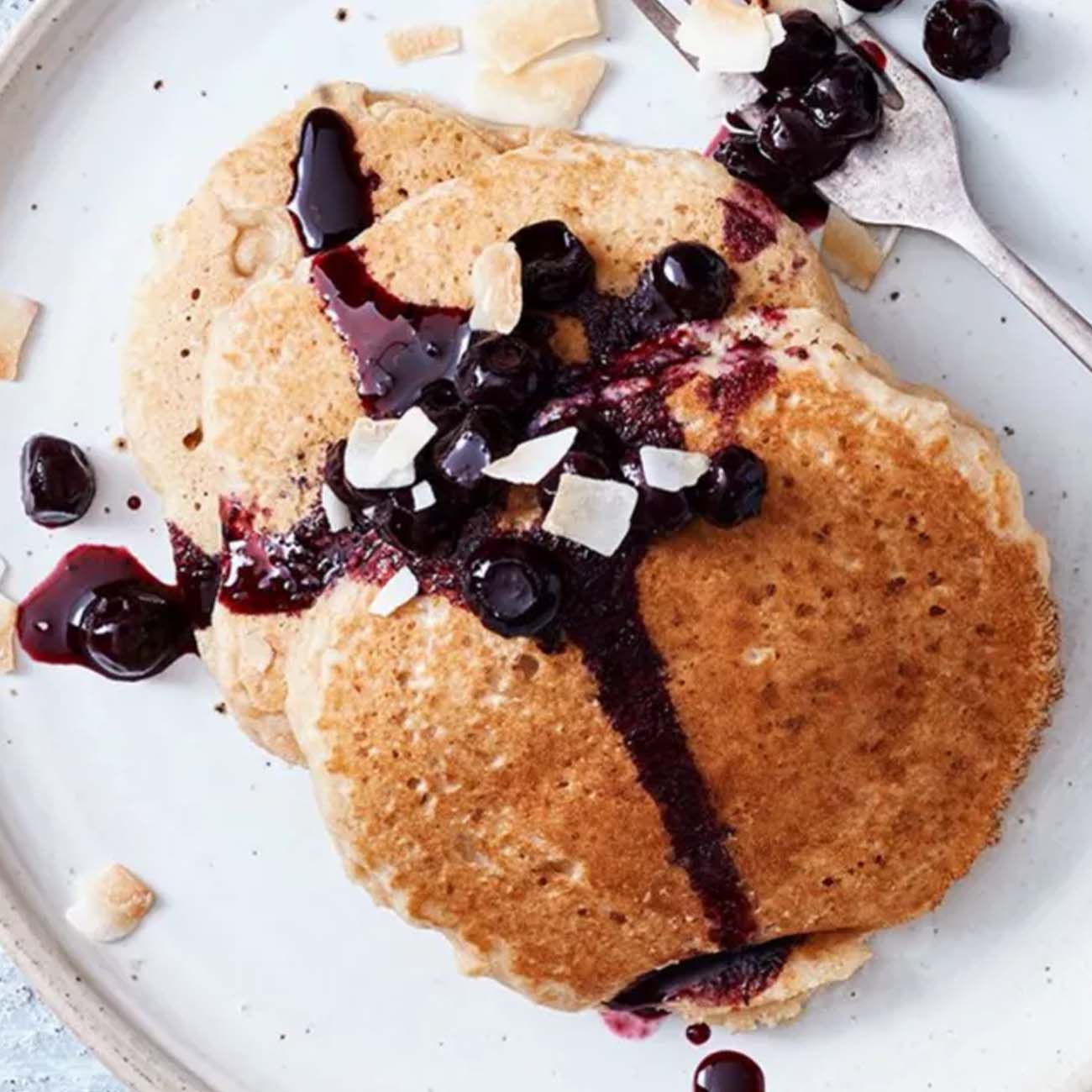A stack of golden brown pancakes on a white plate, topped with a dark berry compote, whole blueberries and coconut flakes.