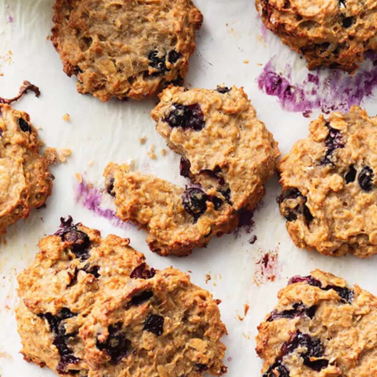 Golden oat and banana cookies packed with whole blueberries, scattered on white baking paper with a few crumbs.