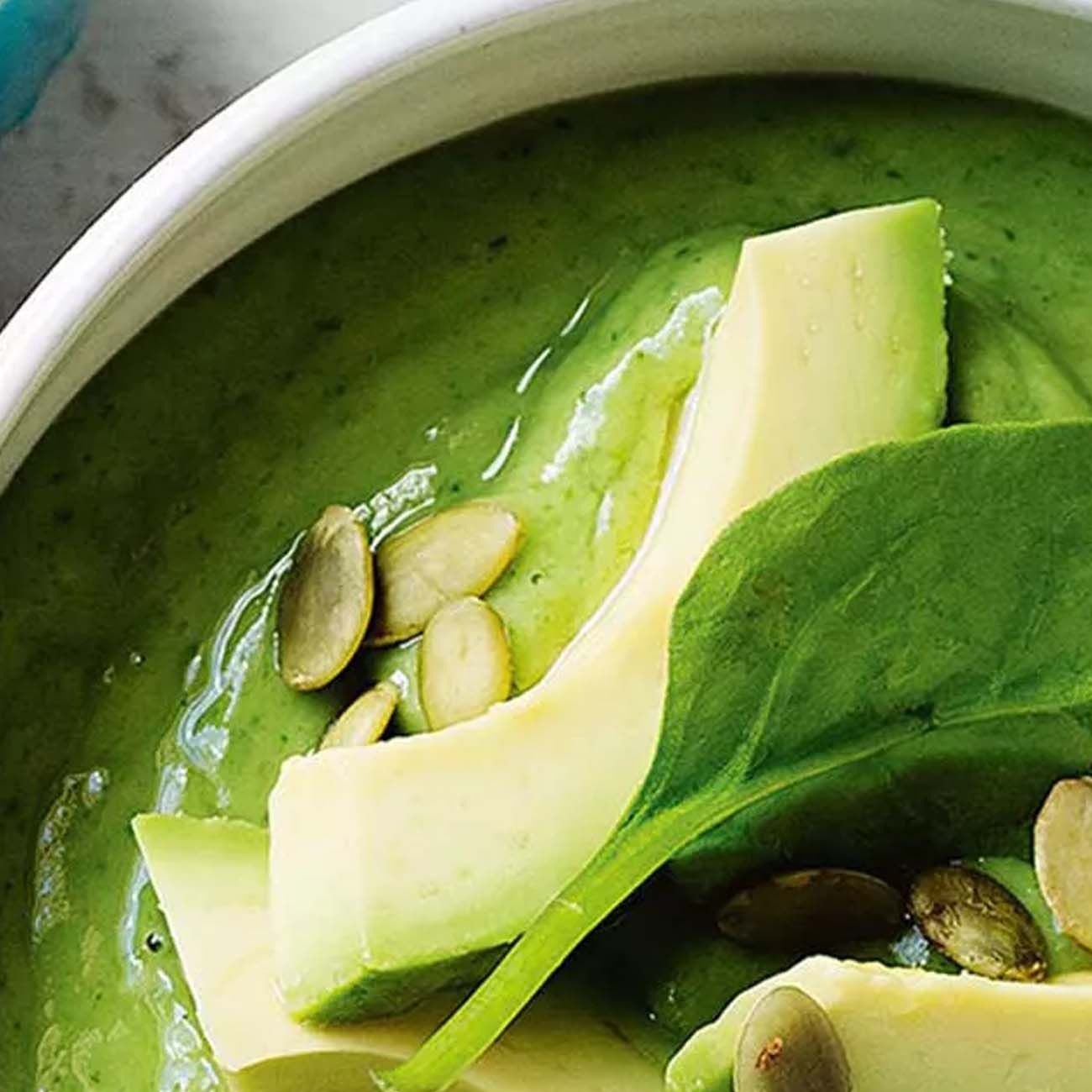 A close-up of a vibrant green smoothie bowl topped with sliced fresh avocado, spinach leaves and pepitas.