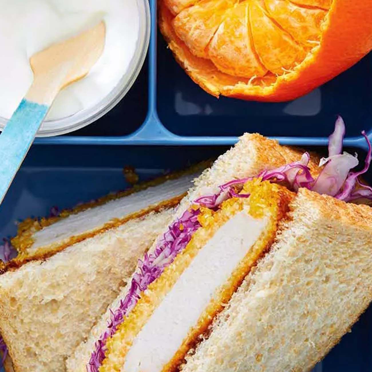 A blue lunchbox compartment containing a schnitzel and red cabbage sandwich, a peeled mandarin and a tub of yoghurt.