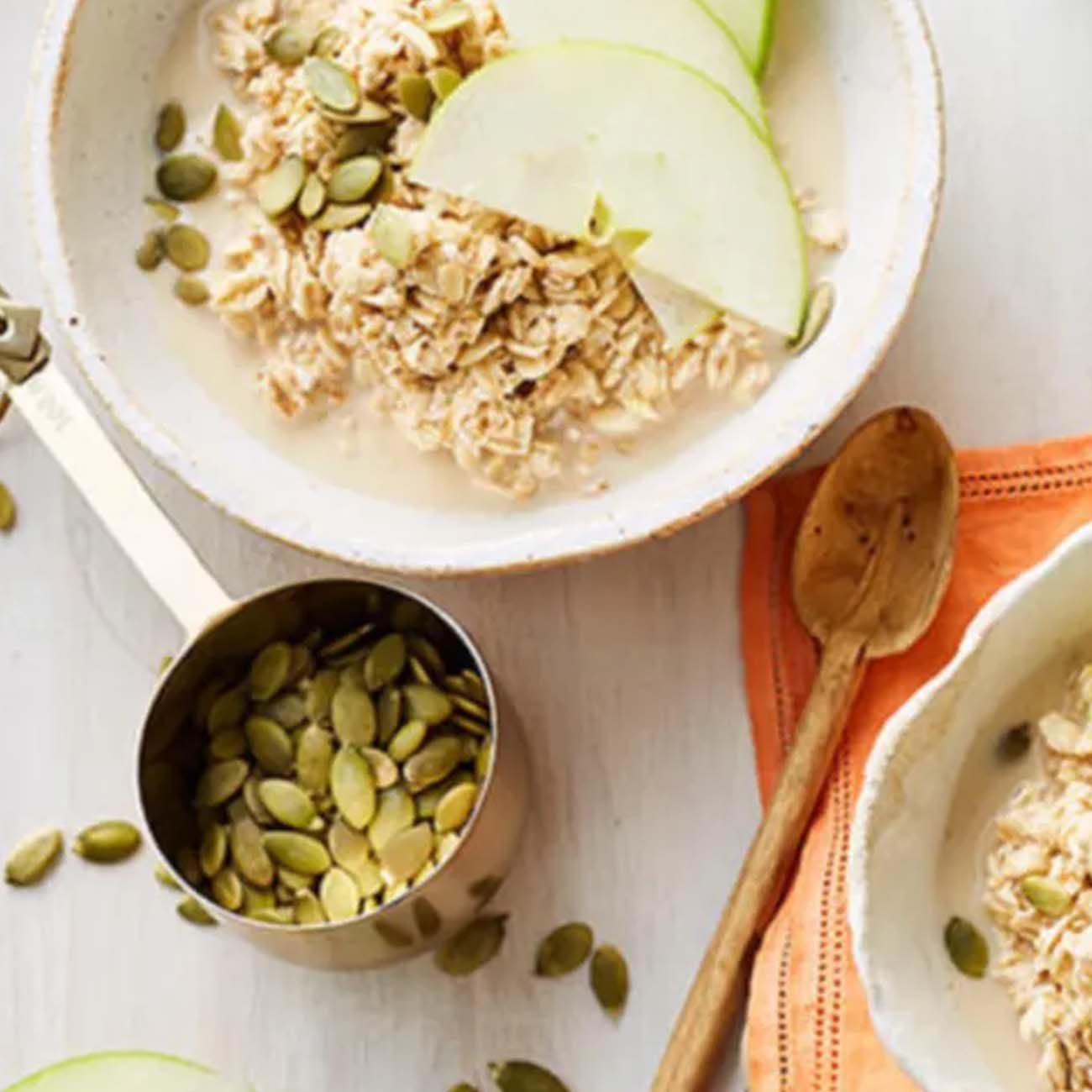 A breakfast bowl of oat porridge topped with green apple slices and pumpkin seeds, next to a measuring scoop of seeds.