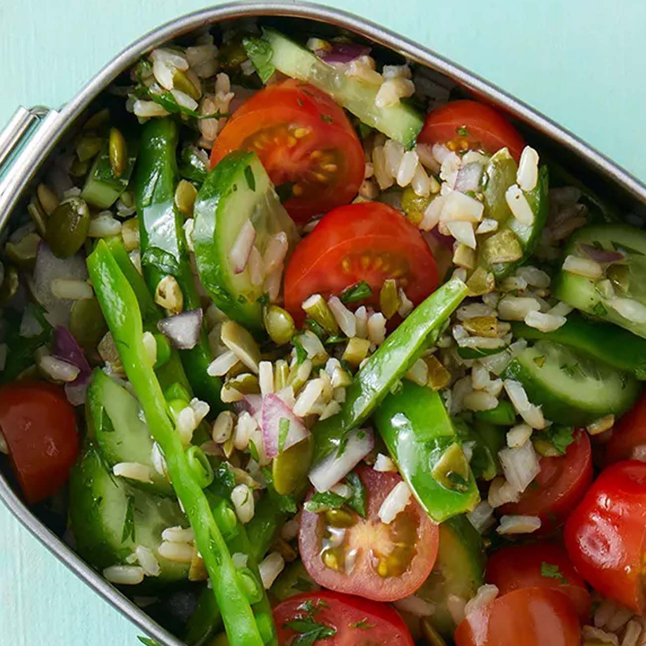 A vibrant rice salad packed with cherry tomatoes, sliced cucumbers, sugar snap peas, red onion and pumpkin seeds in a tin.