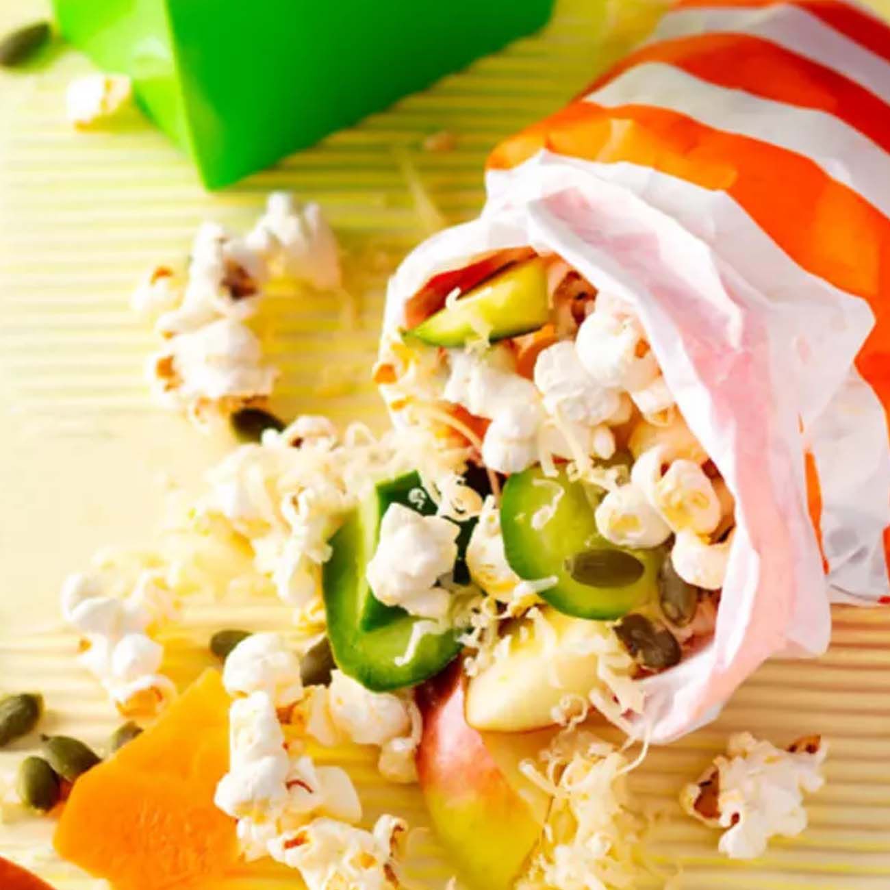 A paper bag spilling popcorn, apple slices, cucumber pieces, grated cheese and pumpkin seeds onto a yellow surface.