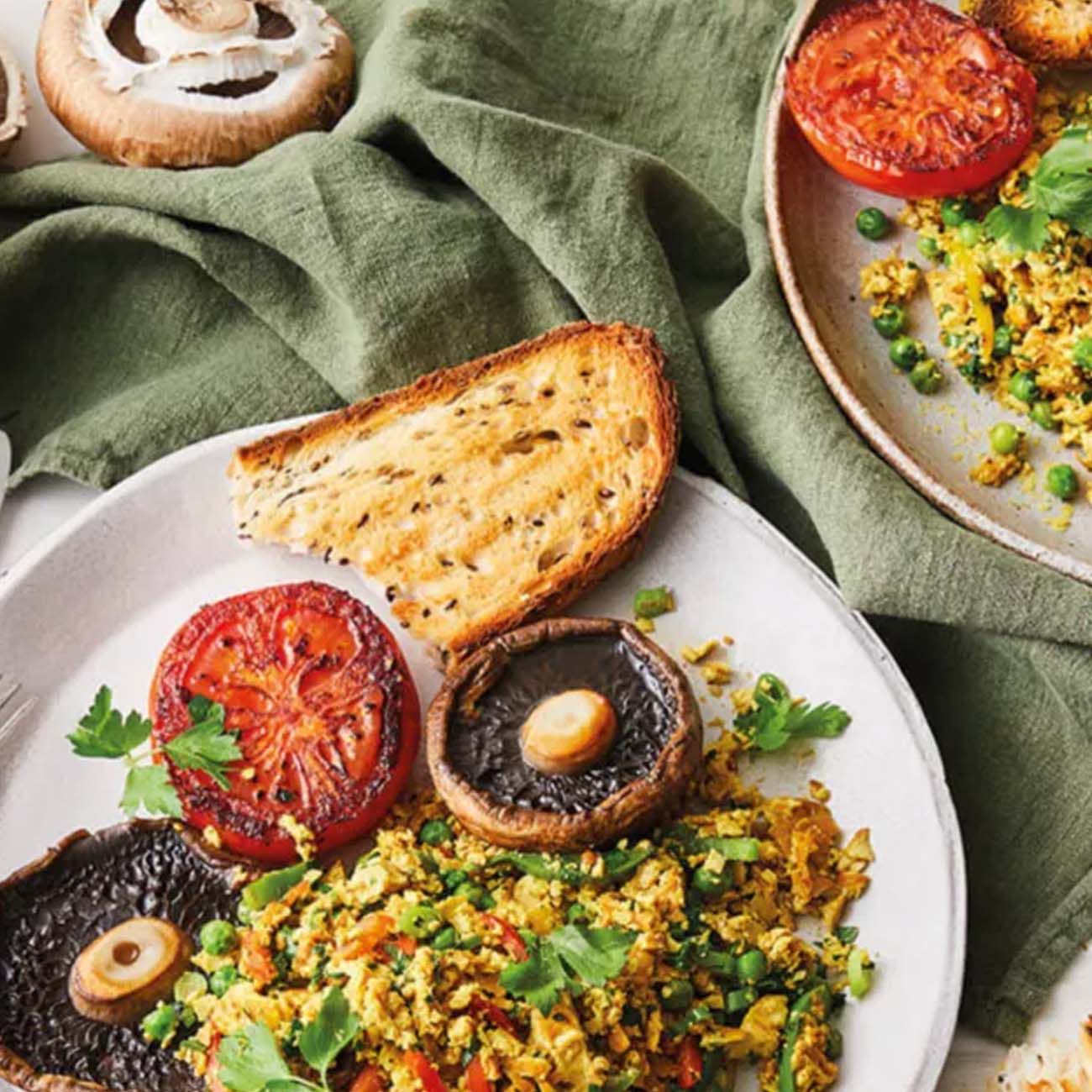 A breakfast plate featuring a tofu or egg scramble with peas, grilled tomatoes, large field mushrooms and toasted bread.