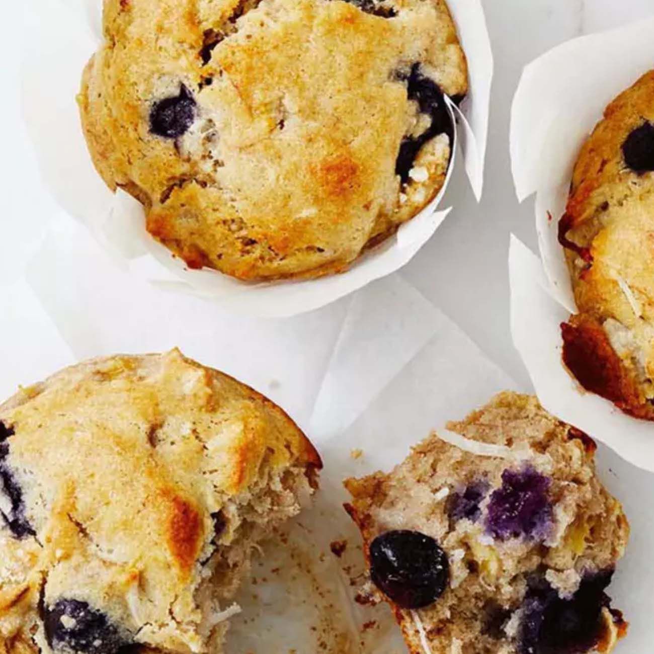 Fluffy blueberry muffins in white paper cases, with one muffin broken open to reveal the soft texture and blue fruit inside.