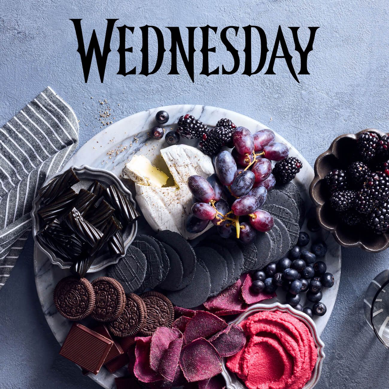 A dark grazing platter with cheese, grapes, and black crackers, featuring the "WEDNESDAY" logo.