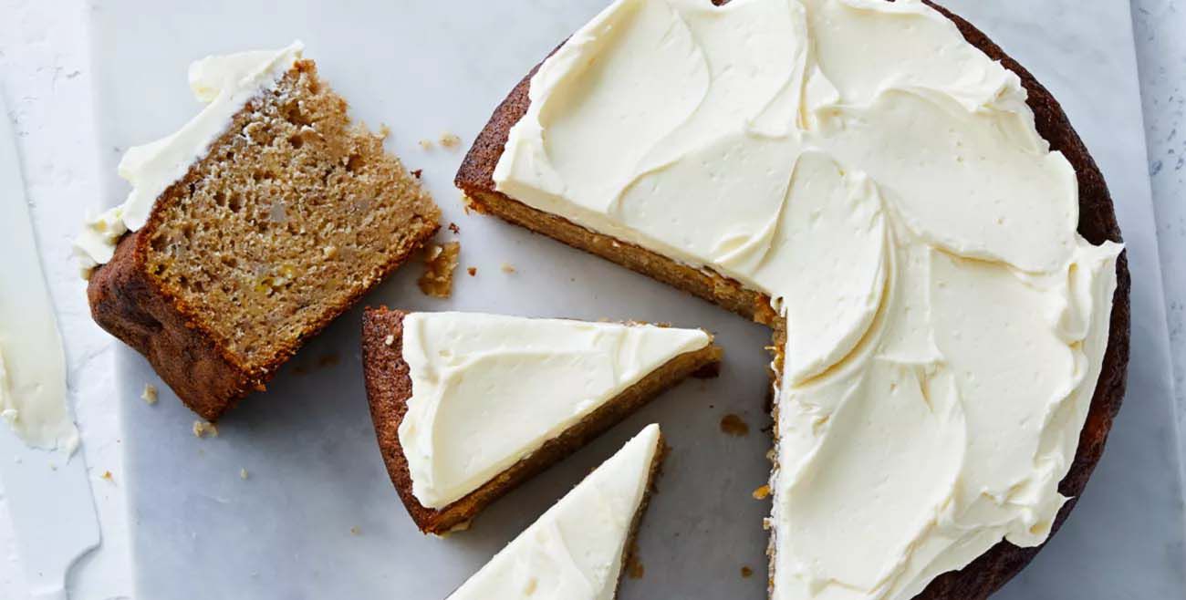 A round banana or carrot cake with thick white frosting, shown cut into slices on a light-coloured marble board.