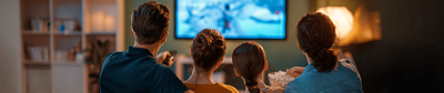 Back view of a family of four sitting on a couch watching a bright TV screen in a dimly lit room with popcorn.
