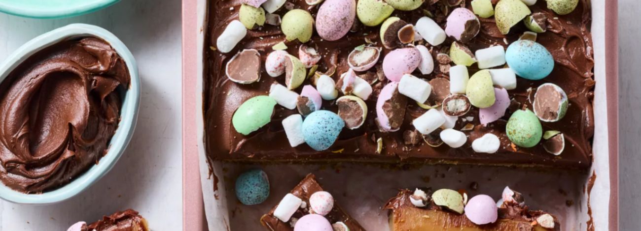 Chocolate caramel slice topped with speckled candy Easter eggs and marshmallows in a baking tin.