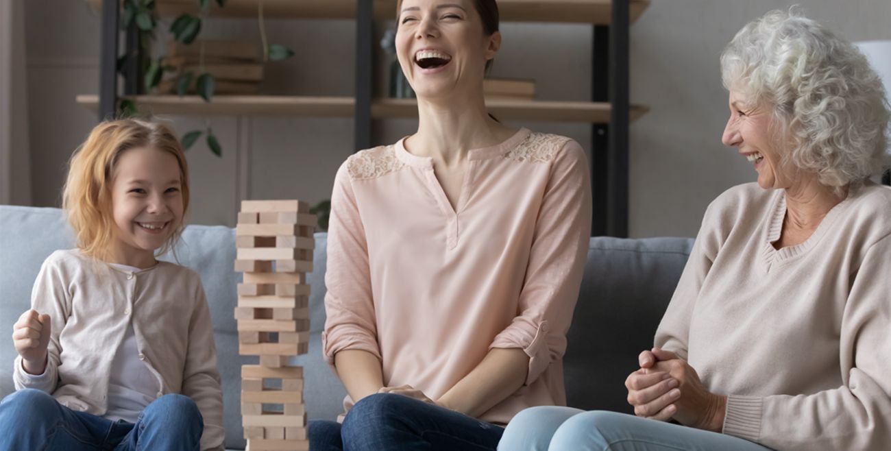 A young girl, woman, and grandmother laugh together while playing a wooden block stacking tower game on a couch.