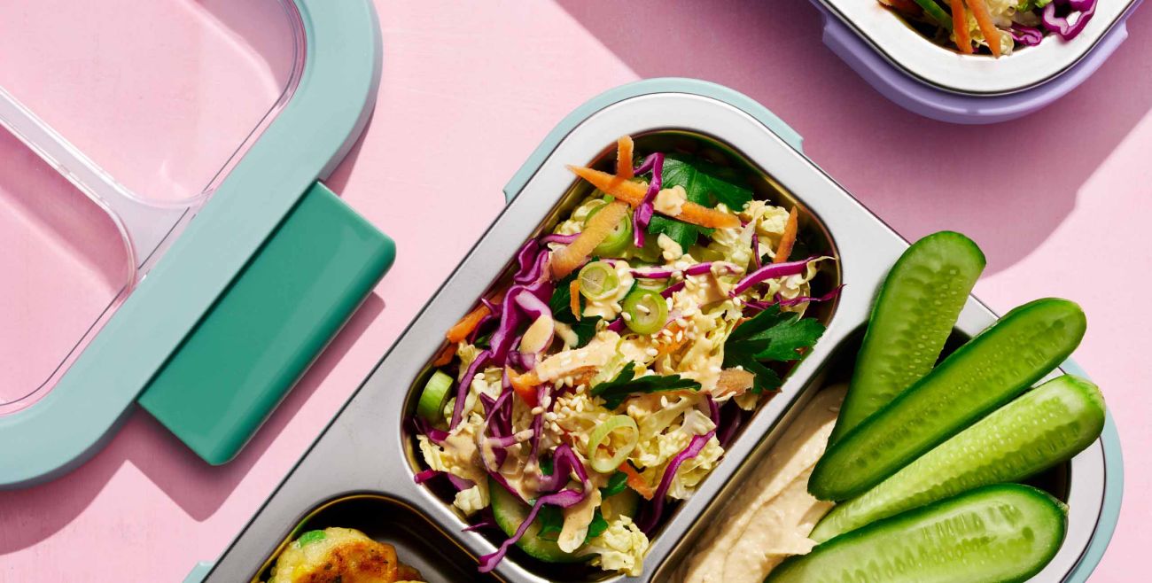 Close-up of a stainless steel bento lunch box packed with cabbage salad, sliced cucumber, and dip