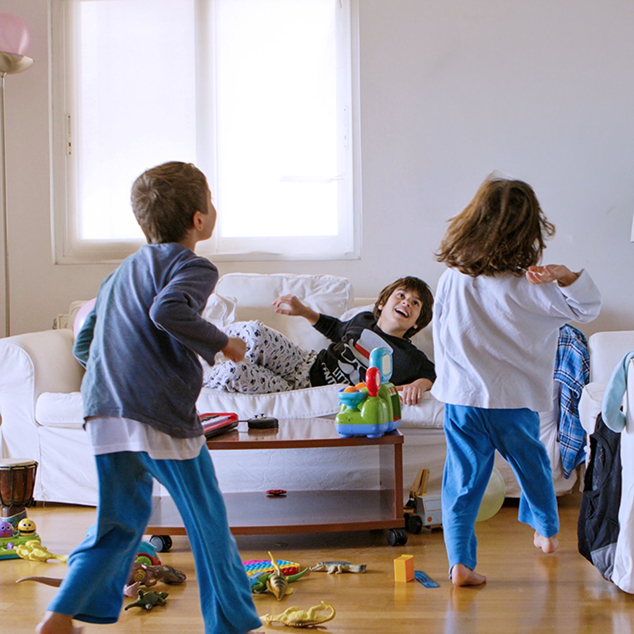Three children in pyjamas play excitedly in a bright living room filled with scattered toys and games.