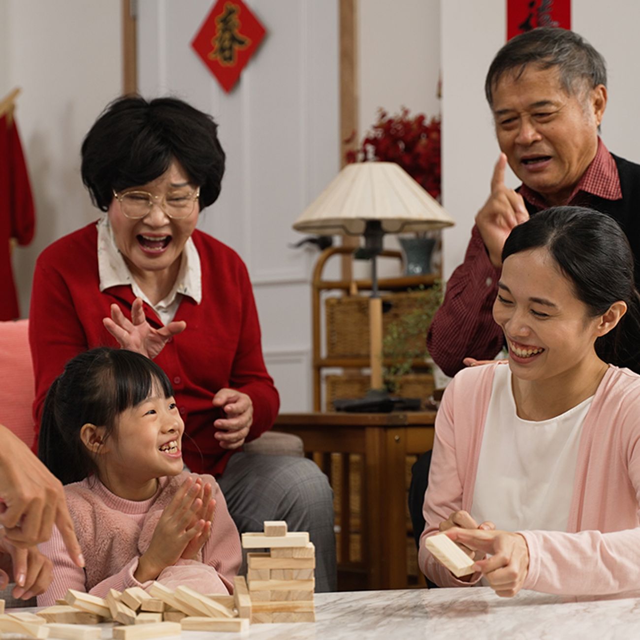 A multi-generational family laughs and cheers while playing a wooden block stacking game in a living room.