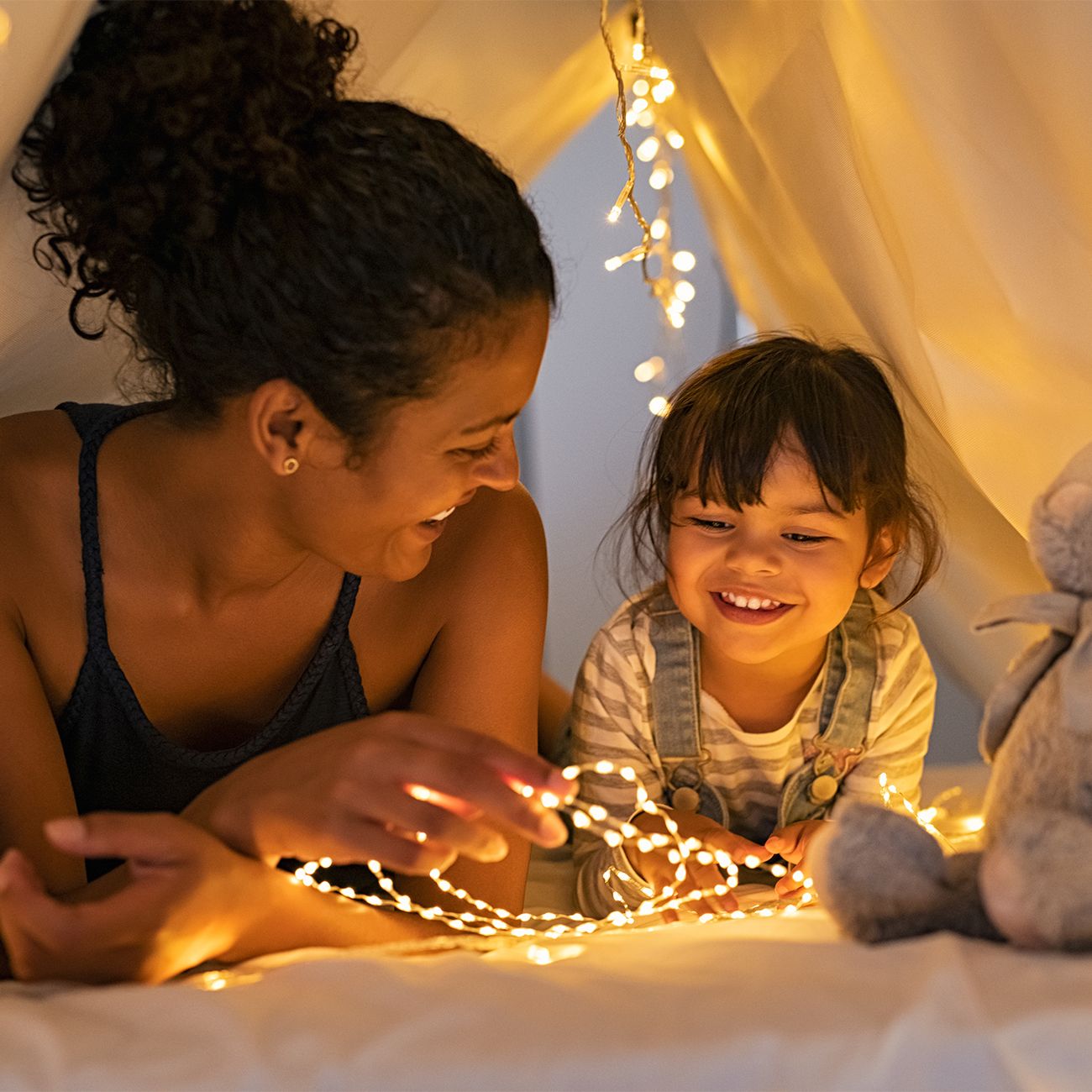 A woman and young girl smile at glowing fairy lights inside a cosy white fabric tent during a home games night.