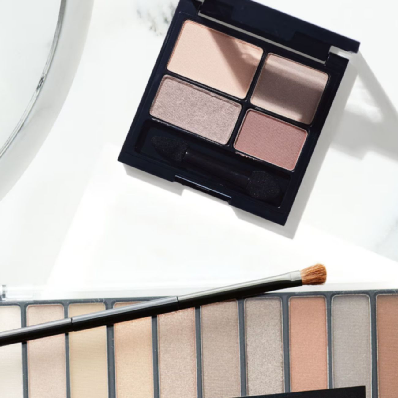 Overhead view of two neutral eyeshadow palettes, a round mirror, and a makeup applicator.