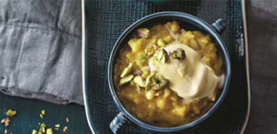 A warm fruit compote served in a blue handled bowl, topped with a dollop of cream and chopped pistachios.