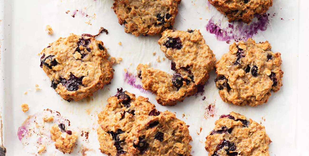 Golden oat and banana cookies packed with whole blueberries, scattered on white baking paper with a few crumbs.