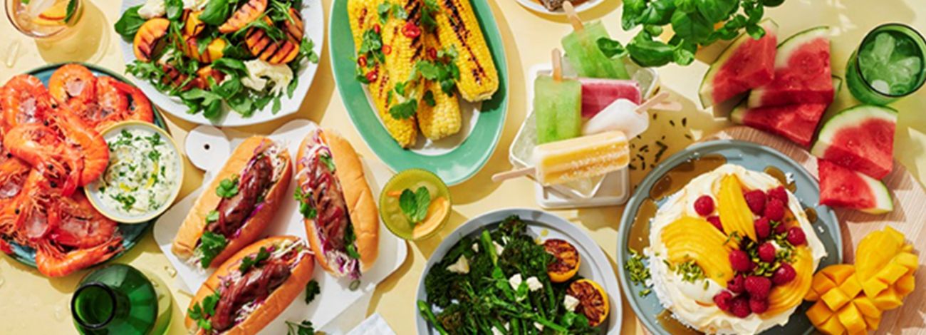 Flat-lay of Australian summer food including grilled prawns, sausages, pavlova, mango, lamingtons and corn cobs.
