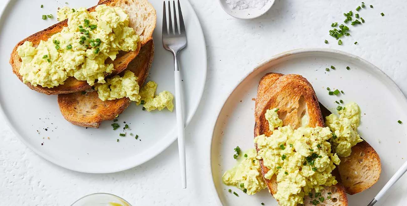 Two plates of creamy tofu scramble on toasted sourdough bread, garnished with fresh chives on a white background.