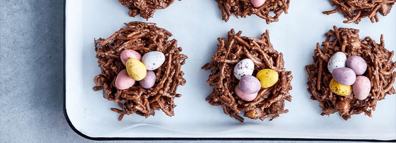 Top-down view of chocolate Easter nests on a white tray, each topped with three pastel-coloured mini eggs.