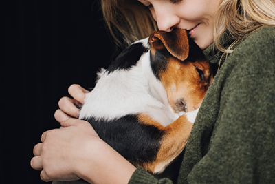 A young woman in a green jumper smiling as she hugs a small, tricolour dog against her chest.